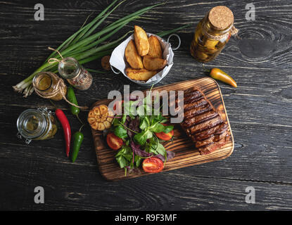 Bistecca su scheda con erbe aromatiche, patate fritte, spezie su una superficie nera Foto Stock