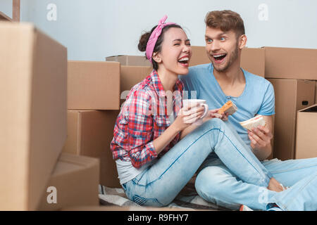 Giovane marito e moglie spostando di nuovo posto seduto sul pavimento afferrando tazze di bere il caffè caldo mangiando panini parlando avendo divertimento ridere per scherzo che Foto Stock