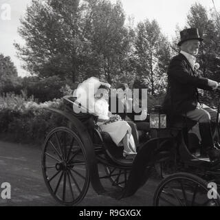 1967, storico, la sposa e lo sposo seduto nella parte posteriore di un antico open-top cavallo pilotato carrello essendo azionato per la ricezione da parte del conducente in calzoncini e indossando un cappello a cilindro, Inghilterra, Regno Unito. Foto Stock
