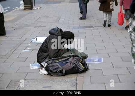 Un senzatetto uomo vestito di nero abbigliamento invernale seduta sul marciapiede marciapiede con testa piegata e la gente a piedi dal London UK Europa KATHY DEWITT Foto Stock