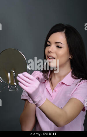 Estetista, cosmetologo su sfondo grigio.un modello con una siringa e uno specchio nelle sue mani fa un iniezione in bocca. Lip augmentation. Foto Stock