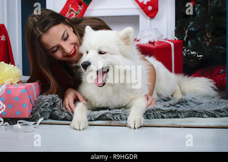 Chiudere ritratto di carino sbadigliare Samoiedo e ragazza sullo sfondo del caminetto bianco e decorazioni di Natale. Cane e il suo proprietario avvolgente Foto Stock