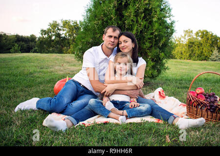 Ritratto di una giovane e bella famiglia felice in un picnic nel parco. Il papà abbraccia la moglie e la figlia Foto Stock