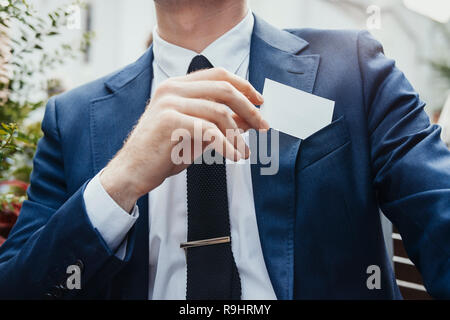 Close up di imprenditore mettendo blank business card nel suo colore blu nella tasca di una giacca. Vista frontale. Foto Stock