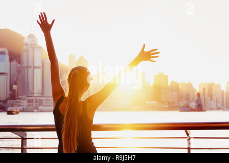 Felice ragazza sta con le mani alzate contro il tramonto e guardando della città di Hong Kong Foto Stock