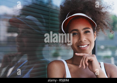 Giovane donna che indossa il tennis visor lo stile libero sulla strada camminare vicino a finestra guardando oltre il pensiero creativo allegro sorridente close-up Foto Stock