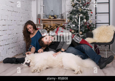 Giovane bella giovane divertirsi con il loro cane sullo sfondo del nuovo anno e decorazioni di Natale. Famiglia con Samoiedo celebrare Foto Stock