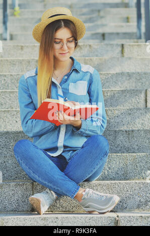 Una ragazza giovane studente vestito in un cappello e una maglietta denim scrive di idee in un notebook e si siede sui passi sulla strada, sorridente verso la telecamera Foto Stock