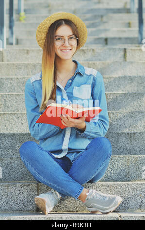 Una ragazza giovane studente vestito in un cappello e una maglietta denim scrive di idee in un notebook e si siede sui passi sulla strada, sorridente verso la telecamera Foto Stock