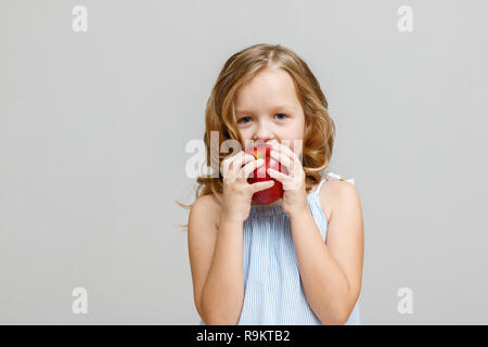 Ritratto di un sorridenti bimba bionda su sfondo grigio. Baby eating red apple Foto Stock