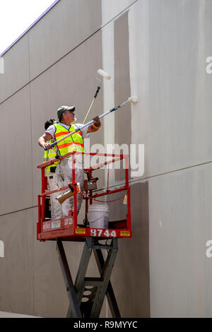 Lavorando da un elevatore a pantografo e sicurezza indossare giubbotti di visibilità, due lavoratori edili utilizzano rulli di vernice dipingere un muro di cemento in Costa Mesa, CA. Foto Stock