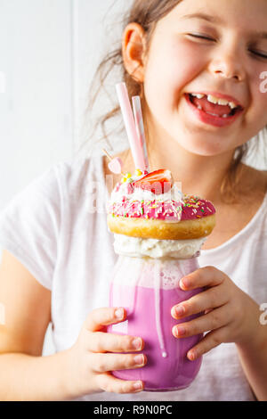 Poco sorridente ragazza bevande Fragola Rosa freak shake cocktail con ciambelle e dolci, sfondo bianco. I dessert malsano concetto. Foto Stock