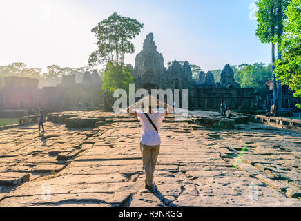 Un turista che visita il tempio Bayon, Angkor Thom rovine, meta di viaggio Cambogia. Donna con cappello tradizionale e bracci sollevati, vista posteriore, sunrise sunb Foto Stock