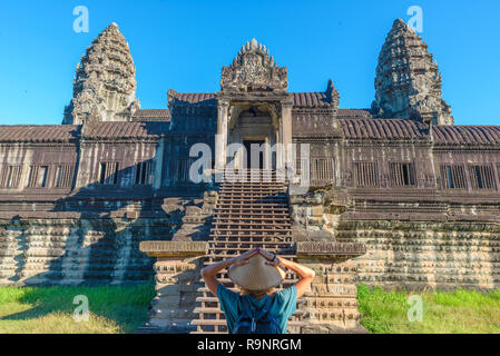 Un turista in visita a Angkor Wat rovine presso sunrise, meta di viaggio Cambogia. Donna con cappello tradizionale e bracci sollevati, vista posteriore, facciata principale stai Foto Stock