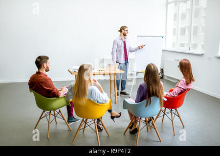 Giovane uomo come un altoparlante reporting per il pubblico durante la riunione in una sala conferenze con colorate sedie Foto Stock
