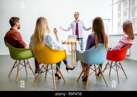 Giovane uomo come un altoparlante reporting per il pubblico durante la riunione in una sala conferenze con colorate sedie Foto Stock