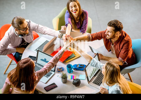 Il team di giovani collaboratori mettendo le mani insieme seduti a tavola rotonda durante il lavoro in ufficio Foto Stock