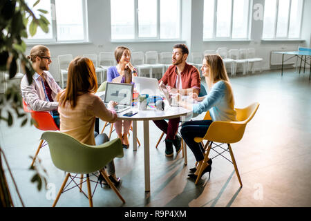 Il team di giovani collaboratori vestito casualmente a lavorare insieme con i laptop seduti al tavolo rotondo in ufficio Foto Stock