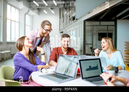 Il team di giovani collaboratori vestito casualmente a lavorare insieme con i laptop seduti al tavolo rotondo in ufficio Foto Stock