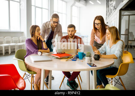 Il team di giovani collaboratori vestito casualmente a lavorare insieme con i laptop seduti al tavolo rotondo in ufficio Foto Stock