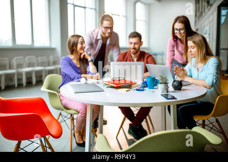 Il team di giovani collaboratori vestito casualmente a lavorare insieme con i laptop seduti al tavolo rotondo in ufficio Foto Stock