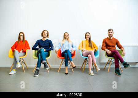 Ritratto di un giovane gente vestita casualmente seduto in una riga sulle sedie colorate sul muro bianco sullo sfondo Foto Stock