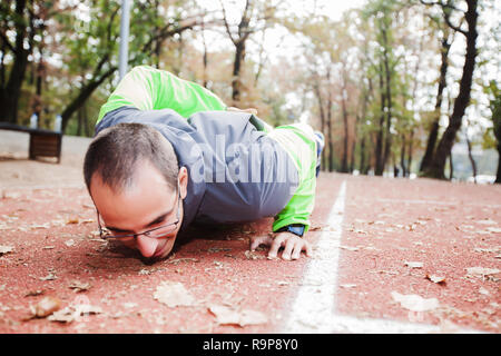 Giovane uomo facendo push-up con una mano, con gli occhiali, in outdoor sports park in autunno la mattina. Uno stile di vita sano allenamento. Foto Stock