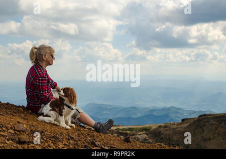 La donna e il suo cane, Cavalier King Charles Spaniel, vestito in royal Stewart, sta guardando il pittoresco paesaggio di montagna Foto Stock