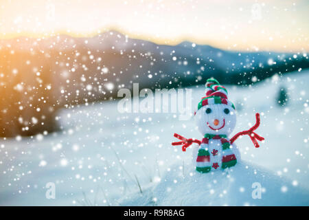 Piccolo giocattolo divertenti baby pupazzo di neve in berretto lavorato a maglia e sciarpa nella neve profonda outdoor sulle montagne sfocata del paesaggio e per la caduta di grossi fiocchi di neve sullo sfondo. Felice Foto Stock