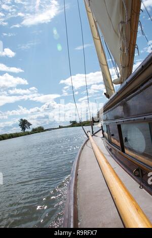 Guardando lungo il lato del ponte in legno tradizionali yacht a vela su un fiume in il Parco Nazionale Broads del Norfolk, Inghilterra. È ventoso e la barca sbandata un acceso Foto Stock