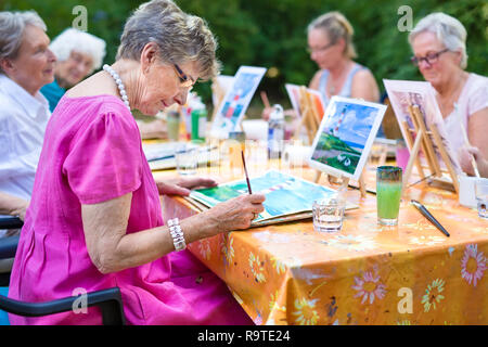 Elegante lady senior pittura in tecnica di classe con gli amici dalla sua casa di cura per gli anziani la copia di un dipinto con acqua di colori su una tela di canapa in esterno ad un Foto Stock