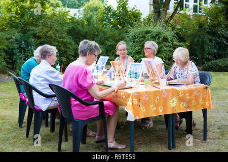 Senior ladies frequentando lezioni di arte all'aperto seduti attorno ad un tavolo a lavorare sul loro dipinti utilizzando il modello Foto su cavalletti. Foto Stock