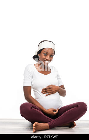 Felice americana africana incinta Donna seduta sul pavimento e mantenendo le mani sulla pancia isolato su bianco Foto Stock