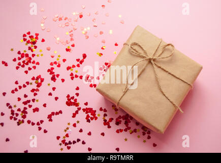 Confezione regalo avvolto in colore marrone carta craft e legato con la fune su sfondo rosa a forma di cuore rosso coriandoli. Foto Stock