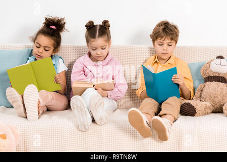 Multietnica adorabili bambini seduti sul divano e la lettura di libri insieme Foto Stock