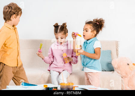 Carino multietnica bambini seduti sul divano e soffiare bolle di sapone insieme Foto Stock