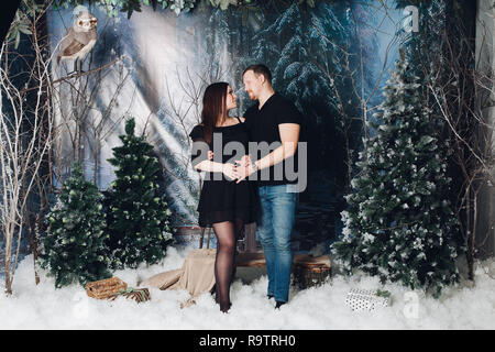 Matura in posa decorate inverno studio con l'abete, alberi di natale e di neve. Uomo con la mano sulla pancia di mia moglie. Bella donna incinta indossa in bl Foto Stock