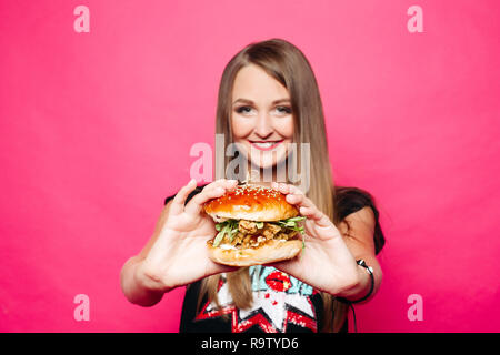 Ritratto in studio di bella donna felice con lunghi capelli biondi holding cheeseburger con pollo e insalata in mani e sorridente in telecamera su rosa backg Foto Stock