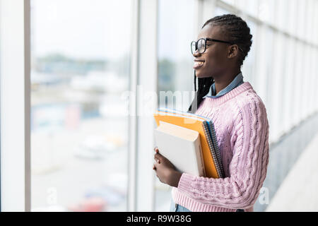 Giovani afro american student donna notebook di contenimento in prossimità di finestre panoramiche nella moderna Università Foto Stock