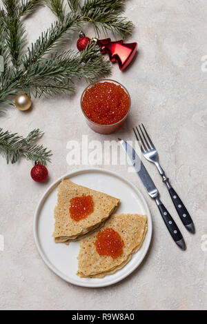 Nuovo anno di prima colazione. Thin gustosi pancake con caviale rosso a piastra bianca con un vasetto di caviale rosso, forcella, coltello, giocattoli di Natale e di abete vicino alla luce bac Foto Stock