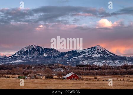 Un tramonto invernale sopra una fattoria lontana e montagne innevate a Delta, Colorado Foto Stock