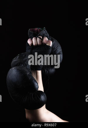 Alzata di mano d'uomo detiene una coppia di vecchi cuoio nero guantoni da pugilato, sfondo nero Foto Stock