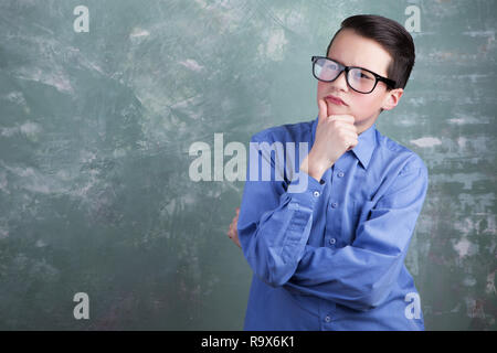 Ritratto di abile young teen boy in bicchieri. Egli è in piedi e guardando pensively e pensare. La sua mano al mento. Sul green grunge background Foto Stock