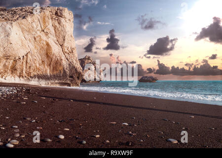 Vista Aprodite rock in Paphos, Cipro con un nuvoloso tramonto e il mare con onde in background. Con sabbia scura e ghiaia e rocce è a Foto Stock