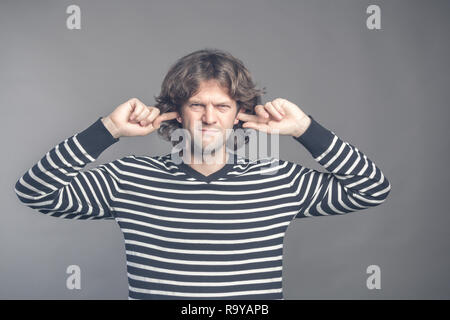 Orinato uomo collegando le orecchie con le dita non vuole ascoltare qualcosa di isolato sul muro grigio Sfondo. Giovane maschio bello di copertura ignorando le orecchie Foto Stock