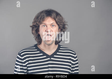 Close up ritratto di funny shaggy pazzo uomo bello in maglione a strisce guardando la fotocamera con grande bocca aperta e la lingua di fuori, teases. Ampia aprì gli occhi Foto Stock