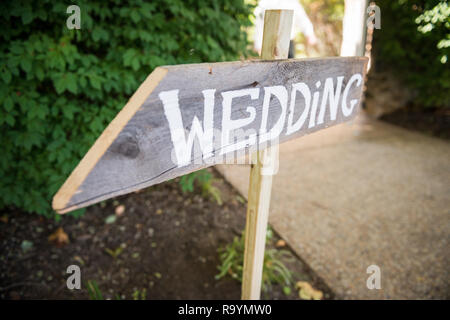 Cartello nozze rustico in legno con scritta bianca su una freccia direzionale che punta verso la cerimonia o il ricevimento. Ambiente esterno con vegetazione. Foto Stock