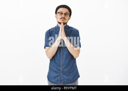 Si prega di miele perdonare me. Ritratto di bello e commovente bello uomo barbuto in bicchieri e nero alla moda beanie tenendo le mani a pregare nei pressi di chin strizzare gli occhi e testa tiltante chiedendo perdono o favore Foto Stock