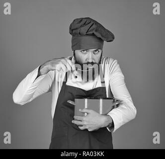 Cuocere con faccia attento in Borgogna uniforme minestra sapori dalla pentola con il cucchiaio. Uomo con barba detiene stoviglie su sfondo rosso. Cucina professionale concetto. Chef con red casseruola e posate Foto Stock