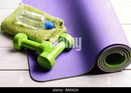 Barbells in prossimità di acqua in bottiglia e asciugamano morbido giacente su viola materassino yoga. Manubri realizzati di plastica verde sulla luce sullo sfondo di legno. La sagomatura e attrezzature per il fitness. Sport e stile di vita sano concetto Foto Stock